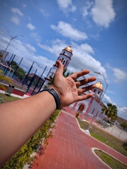 An outstretched hand wearing a black wristband is in the foreground, pointing toward a church or temple with golden domes and white exterior. The scene includes a clear blue sky with clouds and a red-brick walkway leading to the building. In the background, there are trees and a metal fence.