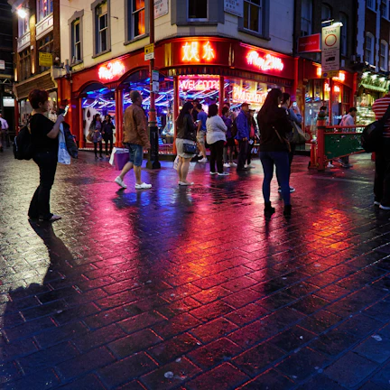 A vibrant street scene at night with glowing neon signs reflecting on wet pavement.