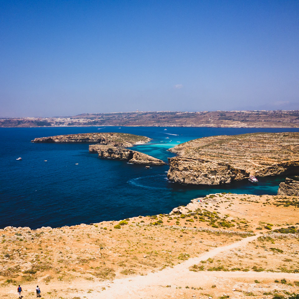 The image depicts a picturesque coastal landscape with two individuals standing on a dirt path that winds through the scene, leading towards the ocean. The sky above them is a clear blue, providing a beautiful backdrop to the natural beauty below. In the distance, several boats can be seen floating on the water, adding to the overall charm of the scene.

The landscape features a variety of rocks and cliffs, with some appearing brown in color due to their age or weathering. The ocean is filled with boats, suggesting that this area might be popular for recreational activities such as sailing or boating.