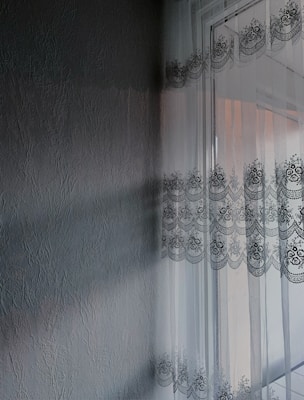 A textured wall with a subtle, embossed pattern is adjacent to a window adorned with sheer lace curtains featuring intricate floral designs. Sunlight filters through the curtains, casting delicate shadows and patterns on the wall.