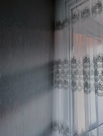 A textured wall with a subtle, embossed pattern is adjacent to a window adorned with sheer lace curtains featuring intricate floral designs. Sunlight filters through the curtains, casting delicate shadows and patterns on the wall.