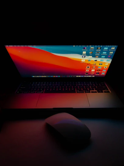 A laptop with a brightly illuminated screen displaying a colorful desktop background and multiple application icons in a dark environment. A wireless mouse is positioned in front of the laptop on a dimly lit surface.