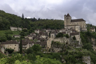 A picturesque medieval village nestled on a hillside, featuring stone houses with brown tiled roofs and a prominent church. The village is surrounded by lush green trees and vegetation, set against an overcast sky.