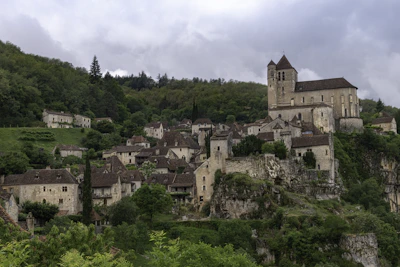 A picturesque medieval village nestled on a hillside, featuring stone houses with brown tiled roofs and a prominent church. The village is surrounded by lush green trees and vegetation, set against an overcast sky.