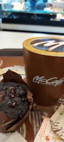 A rich chocolate muffin wrapped in brown paper is positioned next to a tall brown coffee cup with a latte inside, showcasing an intricate design on its frothy top. Both items are placed on a tray with a paper napkin and sugar packet nearby. The background is blurred, displaying the interior of a café.