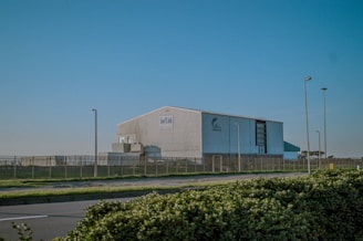 A large industrial building surrounded by a metal fence, situated near a roadway with streetlights. The structure appears to be a warehouse or factory, with clear skies in the background. Shrubbery lines the foreground of the image.