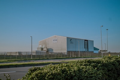 A large industrial building surrounded by a metal fence, situated near a roadway with streetlights. The structure appears to be a warehouse or factory, with clear skies in the background. Shrubbery lines the foreground of the image.