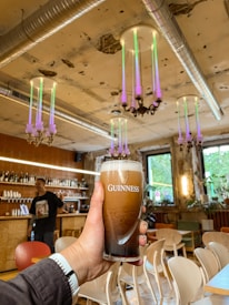A person holds a pint of Guinness beer in a trendy, rustic bar with exposed pipes and colorful hanging lights. The room features wooden furniture and vibrant decor, creating a casual and inviting atmosphere.