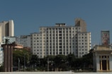 A cityscape featuring a tall, white rectangular building with multiple windows. Surrounding it are various other structures, some of which have graffiti on them. The sky above is clear blue, and there are green trees and a small park area near the base of the buildings. Architectural elements in the foreground include a historic building with intricate designs.