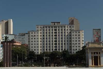 A cityscape featuring a tall, white rectangular building with multiple windows. Surrounding it are various other structures, some of which have graffiti on them. The sky above is clear blue, and there are green trees and a small park area near the base of the buildings. Architectural elements in the foreground include a historic building with intricate designs.