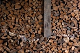 A large pile of chopped firewood neatly stacked, showing the cross sections of various sizes of logs. The image contains a mix of different wood types, colors, and textures. A vertical wooden plank divides the stack slightly off center.