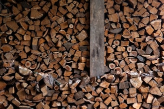 A large pile of chopped firewood neatly stacked, showing the cross sections of various sizes of logs. The image contains a mix of different wood types, colors, and textures. A vertical wooden plank divides the stack slightly off center.