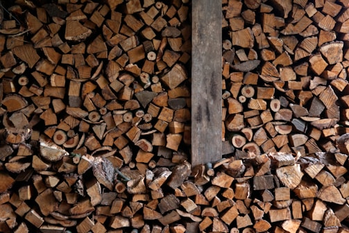 A large pile of chopped firewood neatly stacked, showing the cross sections of various sizes of logs. The image contains a mix of different wood types, colors, and textures. A vertical wooden plank divides the stack slightly off center.