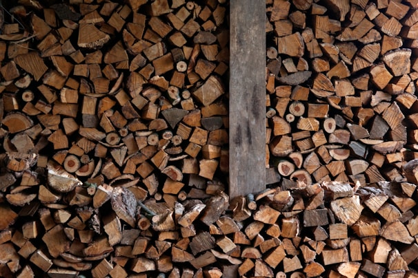 A large pile of chopped firewood neatly stacked, showing the cross sections of various sizes of logs. The image contains a mix of different wood types, colors, and textures. A vertical wooden plank divides the stack slightly off center.