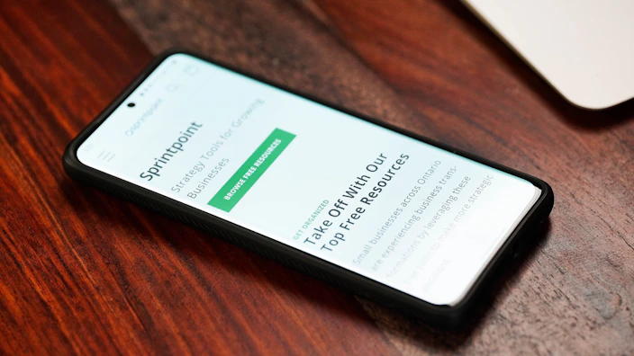 A smartphone with a black case is lying on a wooden surface, displaying a webpage titled 'SprintPoint'. The text on the screen mentions strategic tools for growing businesses and offers free resources.