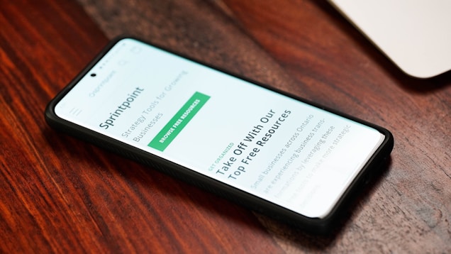 A smartphone with a black case is lying on a wooden surface, displaying a webpage titled 'SprintPoint'. The text on the screen mentions strategic tools for growing businesses and offers free resources.