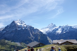 A picturesque alpine landscape featuring two prominent snow-capped mountains under a bright blue sky. Below the mountains, a couple of rustic wooden buildings are situated on a grassy hill. Several people are observing the view, one using binoculars, adding a sense of engagement with the natural scenery.