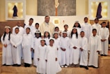 A group of children dressed in white robes, standing together in a church setting with an adult in ceremonial attire. Some of the children are wearing flower crowns. The altar is visible in the background, and there are religious symbols and decorations around.