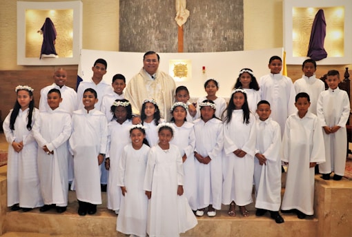 A group of children dressed in white robes, standing together in a church setting with an adult in ceremonial attire. Some of the children are wearing flower crowns. The altar is visible in the background, and there are religious symbols and decorations around.