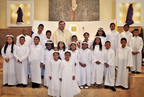 A group of children dressed in white robes, standing together in a church setting with an adult in ceremonial attire. Some of the children are wearing flower crowns. The altar is visible in the background, and there are religious symbols and decorations around.