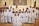 A group of children dressed in white robes, standing together in a church setting with an adult in ceremonial attire. Some of the children are wearing flower crowns. The altar is visible in the background, and there are religious symbols and decorations around.