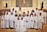 A group of children dressed in white robes, standing together in a church setting with an adult in ceremonial attire. Some of the children are wearing flower crowns. The altar is visible in the background, and there are religious symbols and decorations around.