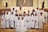 A group of children dressed in white robes, standing together in a church setting with an adult in ceremonial attire. Some of the children are wearing flower crowns. The altar is visible in the background, and there are religious symbols and decorations around.