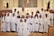 A group of children dressed in white robes, standing together in a church setting with an adult in ceremonial attire. Some of the children are wearing flower crowns. The altar is visible in the background, and there are religious symbols and decorations around.