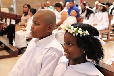 Two children in white robes sit in a church, possibly attending a ceremony. The girl wears a floral crown, while the boy is bald. Behind them, people dressed in white are seated, suggesting a formal or religious event.