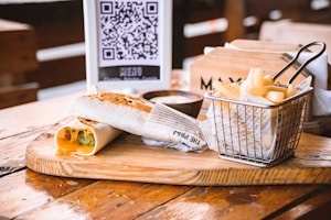 A wooden table holds a serving board with a grilled wrap cut in half, revealing cheese and vegetables inside. Beside it, a small metal basket lined with newspaper and filled with French fries sits next to a small bowl of dipping sauce. In the background, a menu with a QR code is visible next to a wooden box holding napkins.