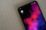 A smartphone with an abstract wallpaper featuring vibrant pink and purple cloud-like patterns on a dark background. The device's screen displays the Facebook Lite app icon, and the status bar shows it is 10:55 with the battery at 55% and connected to Wi-Fi.