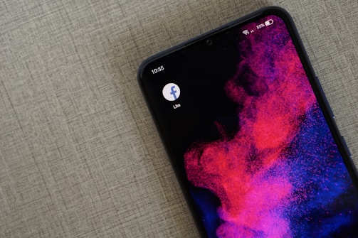 A smartphone with an abstract wallpaper featuring vibrant pink and purple cloud-like patterns on a dark background. The device's screen displays the Facebook Lite app icon, and the status bar shows it is 10:55 with the battery at 55% and connected to Wi-Fi.