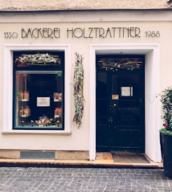 A quaint bakery entrance with decorative foliage and an elegant sign displaying the name 'Bäckerei Holztratnner' along with the numbers 1350 and 1988. The large window displays colorful baked goods and neatly arranged packages. The door and window are framed by a light-colored wall, and the surface in front is paved with cobblestones.