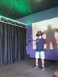 A person is wearing a virtual reality headset, holding controllers, and standing in a room with dark curtains and a poster of a silhouette covered in red paint or blood. The environment appears to be a gaming or simulation space with a technological setup.