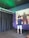 A person is wearing a virtual reality headset, holding controllers, and standing in a room with dark curtains and a poster of a silhouette covered in red paint or blood. The environment appears to be a gaming or simulation space with a technological setup.