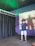 A person is wearing a virtual reality headset, holding controllers, and standing in a room with dark curtains and a poster of a silhouette covered in red paint or blood. The environment appears to be a gaming or simulation space with a technological setup.