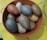A collection of large, oval-shaped stones with a speckled texture is arranged inside a round, brown container with handles. The stones vary in colors such as grey, brown, and beige. The container is resting on a yellow surface.