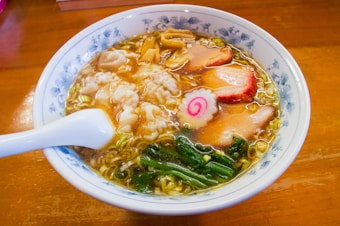 A bowl of ramen with a rich broth, topped with slices of meat, narutomaki, bamboo shoots, and leafy greens. The dish is served in a blue-patterned bowl with a white spoon resting on the edge.