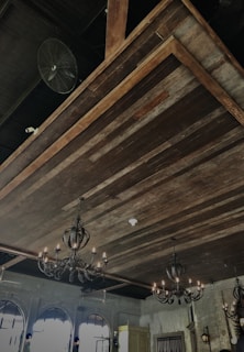 An ornate, rustic interior ceiling design featuring multiple vintage chandeliers with intricate metalwork and lit bulbs. The ceiling is made of wooden planks, giving it a warm and traditional appearance. There are also arched windows and a classic fan attached, adding to the old-fashioned aesthetic. The space exudes an elegant and antique vibe.