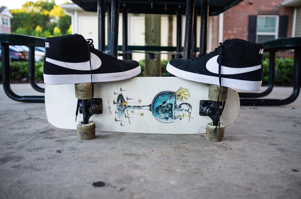 Black high-top sneakers are placed on a skateboard with an artistic graphic design. The skateboard is on a concrete surface beneath a metal picnic table, and there are buildings and greenery in the background.