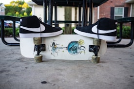 Black high-top sneakers are placed on a skateboard with an artistic graphic design. The skateboard is on a concrete surface beneath a metal picnic table, and there are buildings and greenery in the background.