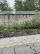 A layered landscape featuring a cracked paved road with yellow lines, a lower stone wall, and a higher rock wall adorned with greenery. Behind the rock wall, there is a metal fence and dense trees in the background under a cloudy sky.