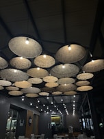 A ceiling design featuring multiple woven basket-like lights with bulbs, creating a rustic and warm ambiance. The lights are arranged in a pattern against a dark industrial ceiling, and below them is a seating area in a modern interior setting.
