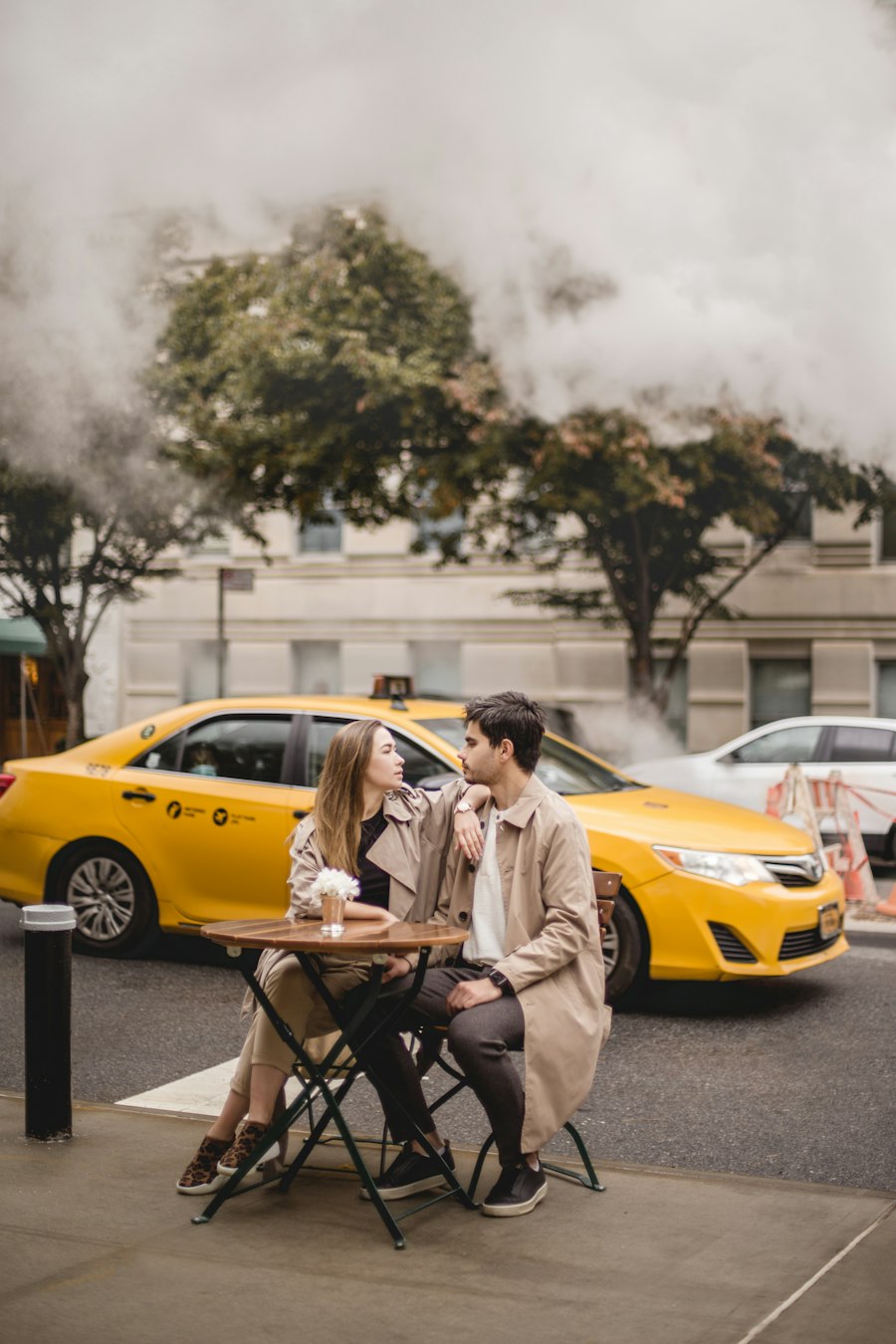 A couple at a café table with a yellow taxi passing