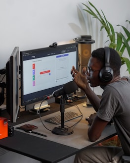 A person wearing headphones is sitting at a desk with a microphone, appearing to communicate or record a podcast. They are using a computer with a monitor displaying a web interface for creating an episode. Other desk items include a keyboard, a desk lamp, and some electronic devices. There's a small potted plant in the background.