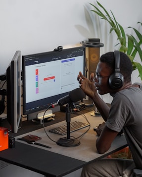 A person wearing headphones is sitting at a desk with a microphone, appearing to communicate or record a podcast. They are using a computer with a monitor displaying a web interface for creating an episode. Other desk items include a keyboard, a desk lamp, and some electronic devices. There's a small potted plant in the background.