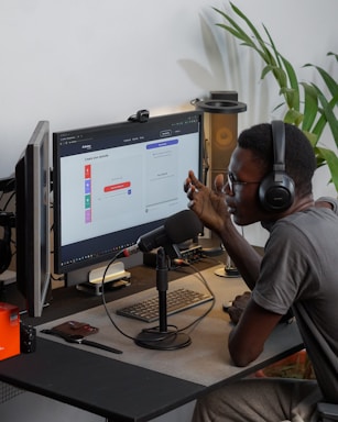A person wearing headphones is sitting at a desk with a microphone, appearing to communicate or record a podcast. They are using a computer with a monitor displaying a web interface for creating an episode. Other desk items include a keyboard, a desk lamp, and some electronic devices. There's a small potted plant in the background.