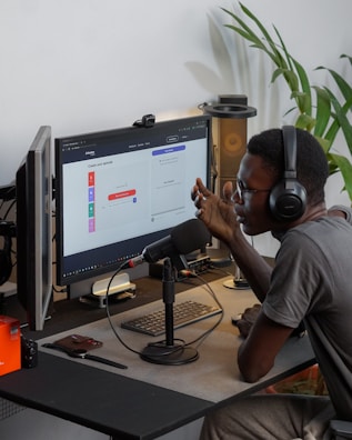 A person wearing headphones is sitting at a desk with a microphone, appearing to communicate or record a podcast. They are using a computer with a monitor displaying a web interface for creating an episode. Other desk items include a keyboard, a desk lamp, and some electronic devices. There's a small potted plant in the background.