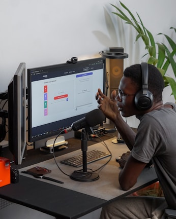 A person wearing headphones is sitting at a desk with a microphone, appearing to communicate or record a podcast. They are using a computer with a monitor displaying a web interface for creating an episode. Other desk items include a keyboard, a desk lamp, and some electronic devices. There's a small potted plant in the background.