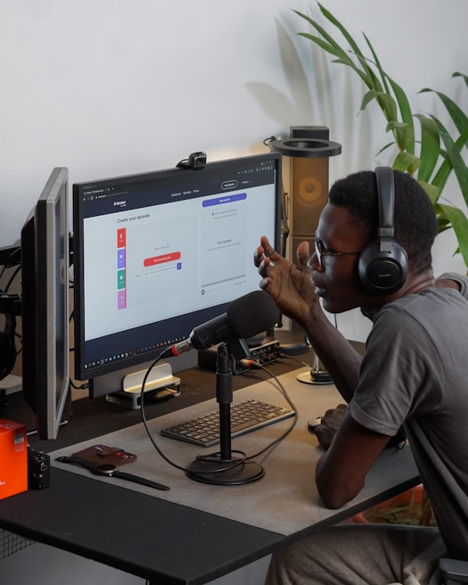 A person wearing headphones is sitting at a desk with a microphone, appearing to communicate or record a podcast. They are using a computer with a monitor displaying a web interface for creating an episode. Other desk items include a keyboard, a desk lamp, and some electronic devices. There's a small potted plant in the background.